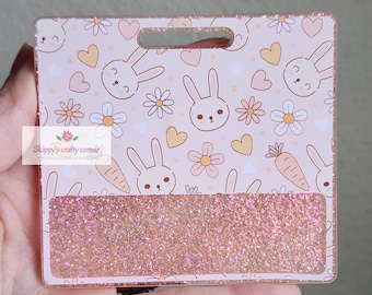 May include: A rectangular acrylic badge holder featuring a light pink background with a repeating pattern of white daisies, orange carrots, small hearts, and cartoon rabbit faces. The bottom section contains a horizontal strip of rose gold glitter.