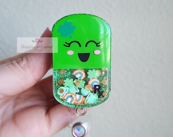 May include: Green resin badge holder shaped like a pill with a smiling face, shamrock, and rainbow glitter inside. The holder has a clear plastic retractable badge clip.