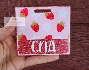Op de afbeelding: Een vierkante acryl badge-topper met een lichtroze achtergrond en een herhalend patroon van rode aardbeien en witte bloemen. Een horizontale rode glitterstrook aan de onderkant toont de witte tekst CNA in een schreefloos lettertype.