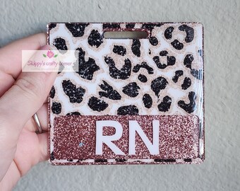 Op de afbeelding: Een vierkante badgehouder met een wit en roségoud glitter luipaardpatroon. Het onderste gedeelte heeft een effen roségouden glitterachtergrond met de witte letters RN in het midden gedrukt. Bovenaan bevindt zich een horizontale gleuf.