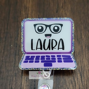 May include: A purple laptop-shaped badge reel with the name "LAURA" in black. The laptop has a smiling face with glasses on the screen and a glittery border. The badge reel has a clear plastic strap and a silver clip with a decorative gem.