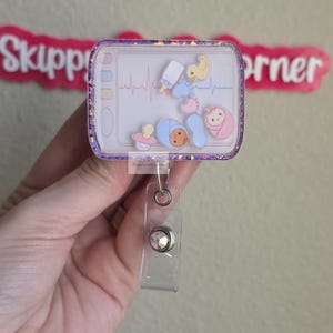 May include: A rectangular badge reel featuring a glittery purple frame and a clear shaker insert. The insert displays a medical heart rate monitor graphic with floating charms including a baby bottle, yellow duck, pacifier, and swaddled infants.