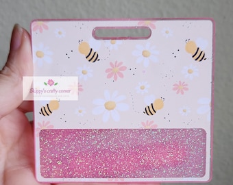 May include: A rectangular badge holder featuring a light beige background with a repeating pattern of yellow and black bees, white daisies, and small pink flowers. The bottom section contains a horizontal rectangular window filled with pink glitter.