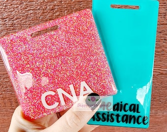 May include: Two rectangular badge holders with horizontal slots at the top. One is a vibrant pink with dense glitter and the white text CNA. The other is a solid teal color with the black text Medical Assistance printed vertically on the surface.