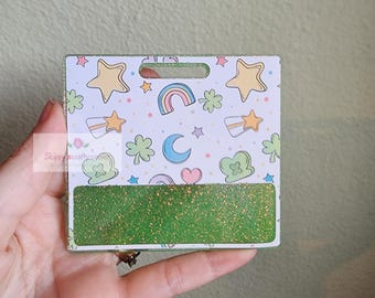 May include: A square badge reel featuring a white background with a repeating pattern of yellow stars, rainbows, blue crescent moons, pink hearts, and green four-leaf clovers. The bottom section has a horizontal band of textured green glitter.