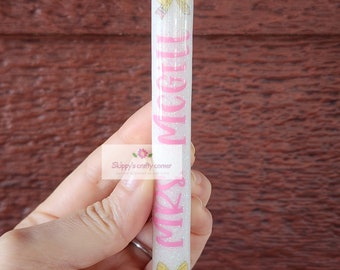 May include: A white glitter pen with a pink "Mrs. McGill" inscription and yellow bows at the top and bottom.