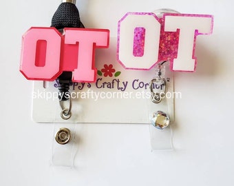 May include: Two badge reels featuring the letters OT. One reel has light pink letters on a dark pink base, attached to a black lanyard. The other reel has white letters on a pink glitter base with a clear strap and a small circular rhinestone.