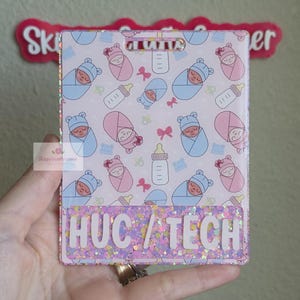 May include: A rectangular badge reel featuring a light pink background with a repeating pattern of swaddled babies in blue and pink, baby bottles, and small bows. The bottom section has a purple glitter finish with the white text HUC / TECH.