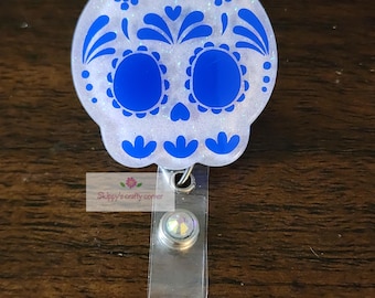 Puede incluir: Un portatarjetas con diseño de calavera blanca y azul del Día de Muertos. El diseño de la calavera presenta patrones florales azules y cuencas de los ojos. El portatarjetas tiene una correa de plástico transparente y un clip plateado. El texto "Skippy's crafty corner" es visible.