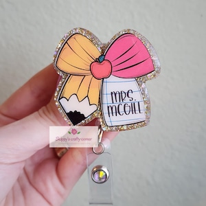 May include: A glittery badge reel featuring a pencil and a pink bow design with an apple accent. The badge reel has the text "MRS. MCGILL" on a notepad. The reel is attached to a clear plastic holder.