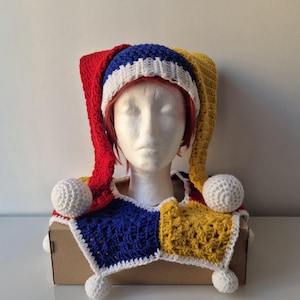 May include: A handmade crochet jester hat and collar set. The hat features red, blue, and yellow panels with white trim and a white pom-pom. The collar mirrors the color scheme with white pom-poms.