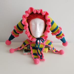 Peut inclure: Un chapeau de bouffon fait à la main au crochet, aux couleurs vives. Le chapeau présente une bordure rose à volants, des sections rayées jaunes et bleues et des pompons roses aux extrémités des longues extensions pointues. Le chapeau est présenté sur une tête de mannequin blanche.