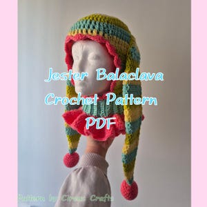 May include: A crochet jester balaclava in a striped pattern of yellow, blue, and green. It has a pink ruffled collar and pom-poms. The image includes the text "Jester Balaclava Crochet Pattern PDF".