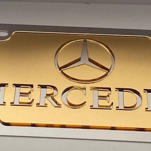 May include: A gold-colored license plate frame with a silver border. The frame features the Mercedes-Benz logo and the word "MERCEDES" in silver lettering.