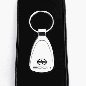 May include: A silver key chain with a black Scion logo and a silver key ring. The key chain is in a black velvet box.