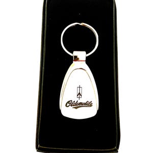 May include: A white Oldsmobile keychain with a silver key ring, presented in a black box. The keychain features the Oldsmobile logo and text in a stylized font.