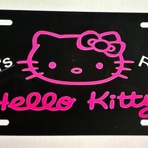 May include: Black license plate featuring a pink Hello Kitty design. The text "Riah's Ride" is in white, and "Hello Kitty" is in pink. The plate has four mounting holes.