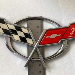 May include: A chrome car emblem with a checkered flag and a red shield with a fleur-de-lis. The emblem is a popular symbol for the Chevrolet Corvette.