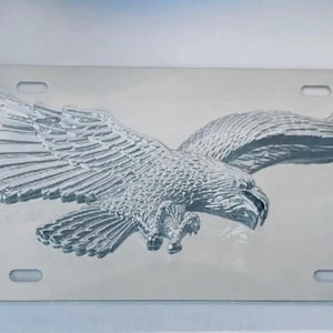 May include: A silver metal license plate featuring a 3D embossed eagle with wings spread wide. The eagle is facing right and has a detailed design.