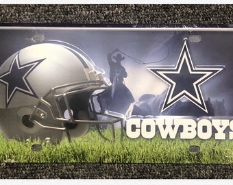 May include: A metal license plate featuring a silver football helmet with a blue star, a silhouette of a cowboy riding a horse, and a blue star with a white outline. The text "COWBOYS" is printed in white letters on a green grass background.
