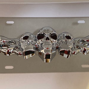 May include: Chrome license plate frame with three skull heads and flames. The skulls have red eyes and are in a row with flames on either side.