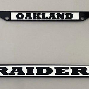 May include: Black license plate frame with the word "OAKLAND" at the top and "RAIDERS" at the bottom in bold, white letters. The frame is rectangular and has a glossy finish.