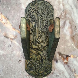May include: A hand-carved wooden wall-mounted shelf with a detailed relief carving. The carving depicts two figures in a forest setting, with intricate details and a weathered, green-toned finish. The shelf has two side supports.
