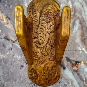 Puede incluir: Un servilletero vertical de madera con una talla en relieve detallada de un elefante rodeado de árboles y follaje. La veta natural de la madera es visible en toda la pieza, que cuenta con dos paneles laterales verticales y una base.