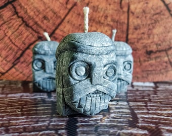 May include: Three gray candle sculptures shaped like human heads with carved facial features. The candles have a rough, textured surface and are lit with white wicks.
