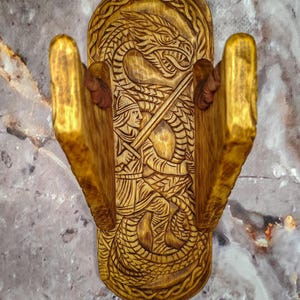 May include: Wooden wall-mounted sword holder with a carved design. The holder features a central panel with a warrior battling a dragon, flanked by two arms to hold the sword. The wood has a warm, golden-brown hue.