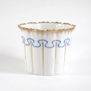 May include: White ceramic planter with a scalloped gold rim and vertical ridges. The planter features a blue ribbon design and gold accents. A decorative container for plants or small items.