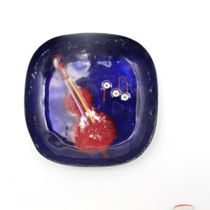 May include: A square, dark blue ceramic dish with a glossy finish. The dish features abstract red, yellow, and white designs. The dish is likely used for decorative purposes or as a small serving piece.
