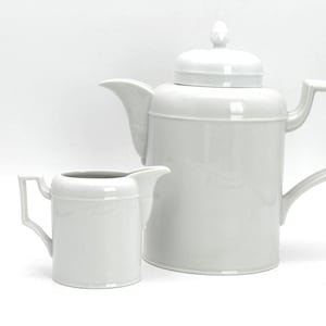 Puede incluir: Un juego de té de cerámica blanca, que incluye una tetera con tapa y una jarra para la crema. La tetera tiene un asa cuadrada y un pico. La jarra de crema tiene un diseño similar. Sobre un fondo blanco.