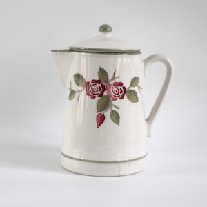Puede incluir: Cafetera de cerámica color crema con tapa y asa. Decorada con un diseño floral de rosas rojas y hojas verdes. El borde y la base tienen un ribete verde. Un artículo de cocina clásico.