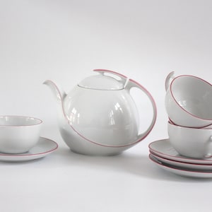 May include: A white teapot and four white teacups with pink trim. The teapot has a unique, curved design. The teacups are stacked on top of each other, with the top cup having a handle.