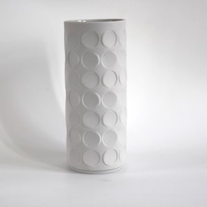 May include: A tall, cylindrical vase in a light gray color. The vase features a repeating pattern of raised, circular designs across its surface. The vase is made of ceramic or a similar material and has a smooth finish.