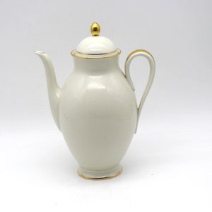 Può includere: Caffettiera in ceramica color crema con coperchio e manico curvo. La caffettiera ha un lungo beccuccio ed è rifinita con finiture dorate sul bordo, sul manico e sulla base. Il coperchio è sormontato da un pomello dorato.