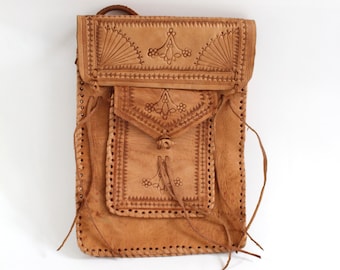 May include: A tan leather shoulder bag with intricate carved floral and geometric designs. The bag features a flap closure, a front pocket, and a long strap. The edges are stitched and the bag has a rustic, handcrafted appearance.