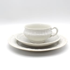 May include: A white teacup and saucer set. The teacup sits atop a small saucer, which rests on a larger plate. All pieces have a textured, ridged design. The set is made of ceramic and is on a white background.