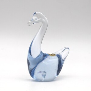May include: A decorative glass swan figurine. The swan is primarily clear with blue accents throughout the body and neck. The figurine has a long, curved neck and a small, detailed head. A small gold sticker is on the body.
