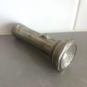 May include: A vintage cylindrical metal flashlight with a weathered silver finish. The body features vertical ridges, a prominent rectangular switch on the side, and a circular head with a glass lens. The metal surface shows signs of wear.