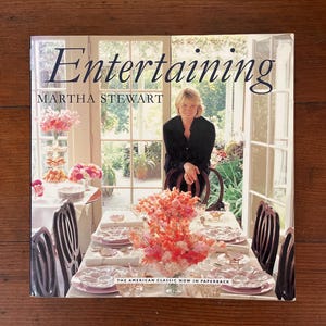 May include: A book titled "Entertaining" by Martha Stewart. The cover features a photo of a woman standing in a dining room with a table set for a meal. The table is decorated with pink flowers and white dishes. The text "The American Classic Now in Paperback" is printed on the bottom of the cover.