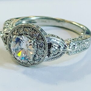 Puede incluir: Un anillo de compromiso de plata con una gran piedra central redonda rodeada de diamantes m&aacute;s peque&ntilde;os. La banda est&aacute; decorada con detalles intrincados y presenta un dise&ntilde;o delicado.