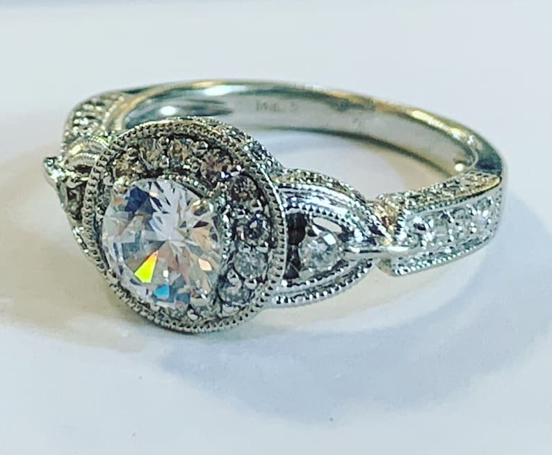 Puede incluir: Un anillo de compromiso de plata con una gran piedra central redonda rodeada de diamantes m&aacute;s peque&ntilde;os. La banda est&aacute; decorada con detalles intrincados y presenta un dise&ntilde;o delicado.
