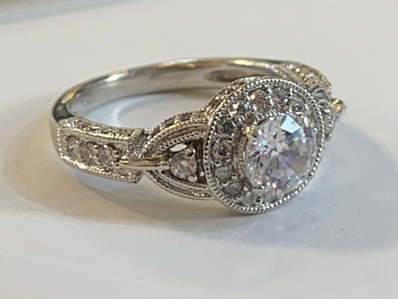 Puede incluir: Un anillo de plata con una gran piedra redonda central rodeada de piedras m&aacute;s peque&ntilde;as. La banda est&aacute; decorada con detalles intrincados.