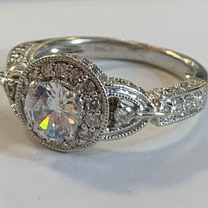 Puede incluir: Un anillo de compromiso de plata con una gran piedra central redonda y clara rodeada de piedras m&aacute;s peque&ntilde;as. La banda est&aacute; decorada con detalles intrincados y presenta un dise&ntilde;o delicado.