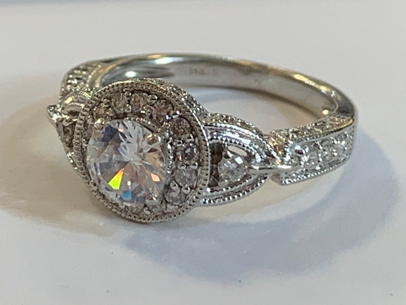 Puede incluir: Un anillo de compromiso de plata con una gran piedra central redonda y clara rodeada de piedras m&aacute;s peque&ntilde;as. La banda est&aacute; decorada con detalles intrincados y presenta un dise&ntilde;o delicado.