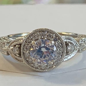 Puede incluir: Un anillo de compromiso de plata con una gran piedra central redonda rodeada de piedras m&aacute;s peque&ntilde;as. La banda est&aacute; intrincadamente dise&ntilde;ada con un delicado patr&oacute;n trenzado.