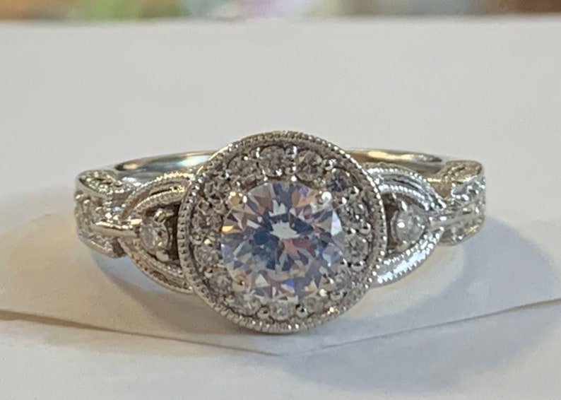 Puede incluir: Un anillo de compromiso de plata con una gran piedra central redonda rodeada de piedras m&aacute;s peque&ntilde;as. La banda est&aacute; intrincadamente dise&ntilde;ada con un delicado patr&oacute;n trenzado.