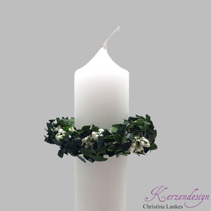 Könnte beinhalten: Eine weiße Stumpenkerze mit einem grünen und weißen Blumenkranz um die Basis. Die Kerze steht auf einem grauen Hintergrund.